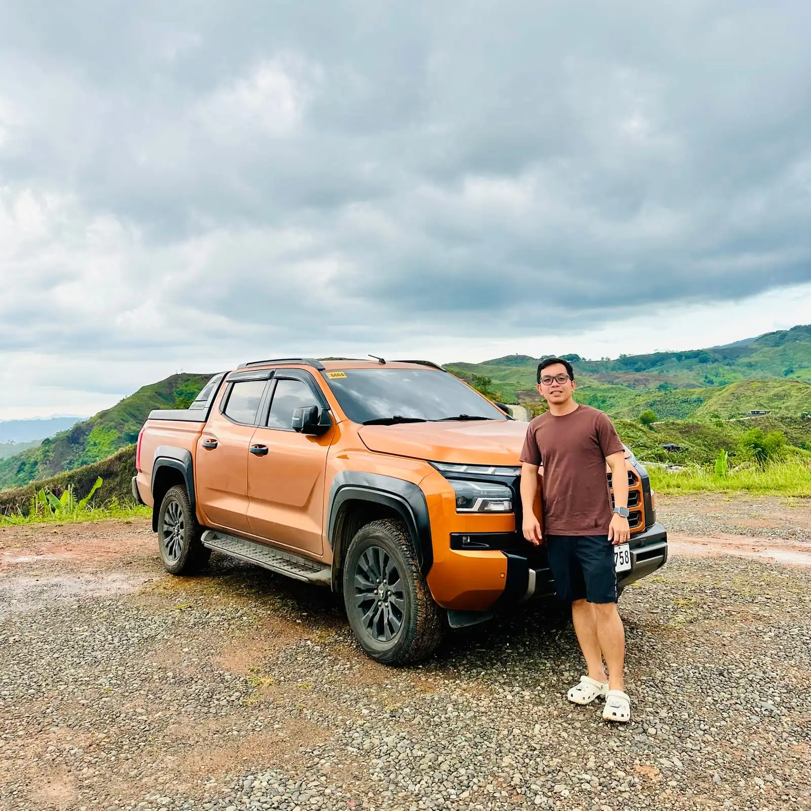 Leaning on an orange pickup truck at a hilltop lookout with green hills in the distance
