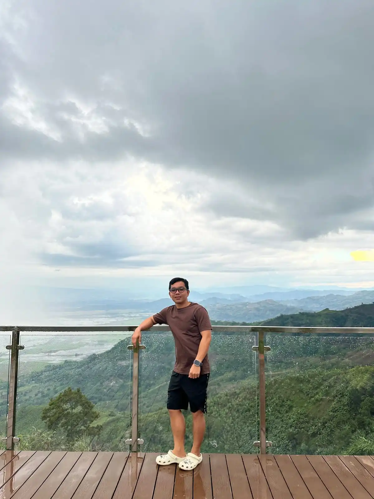 Leaning on a glass railing at a mountain viewing deck under heavy clouds